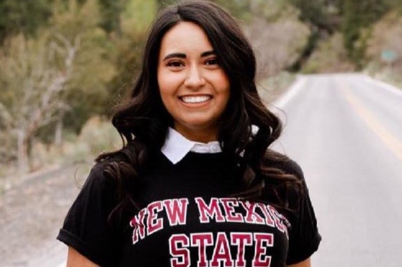 Carleen Silva smiling and wearing a black "New Mexico State" shirt with a white-collared shirt underneath. Coniferous trees and a road leading away from her are in the background.