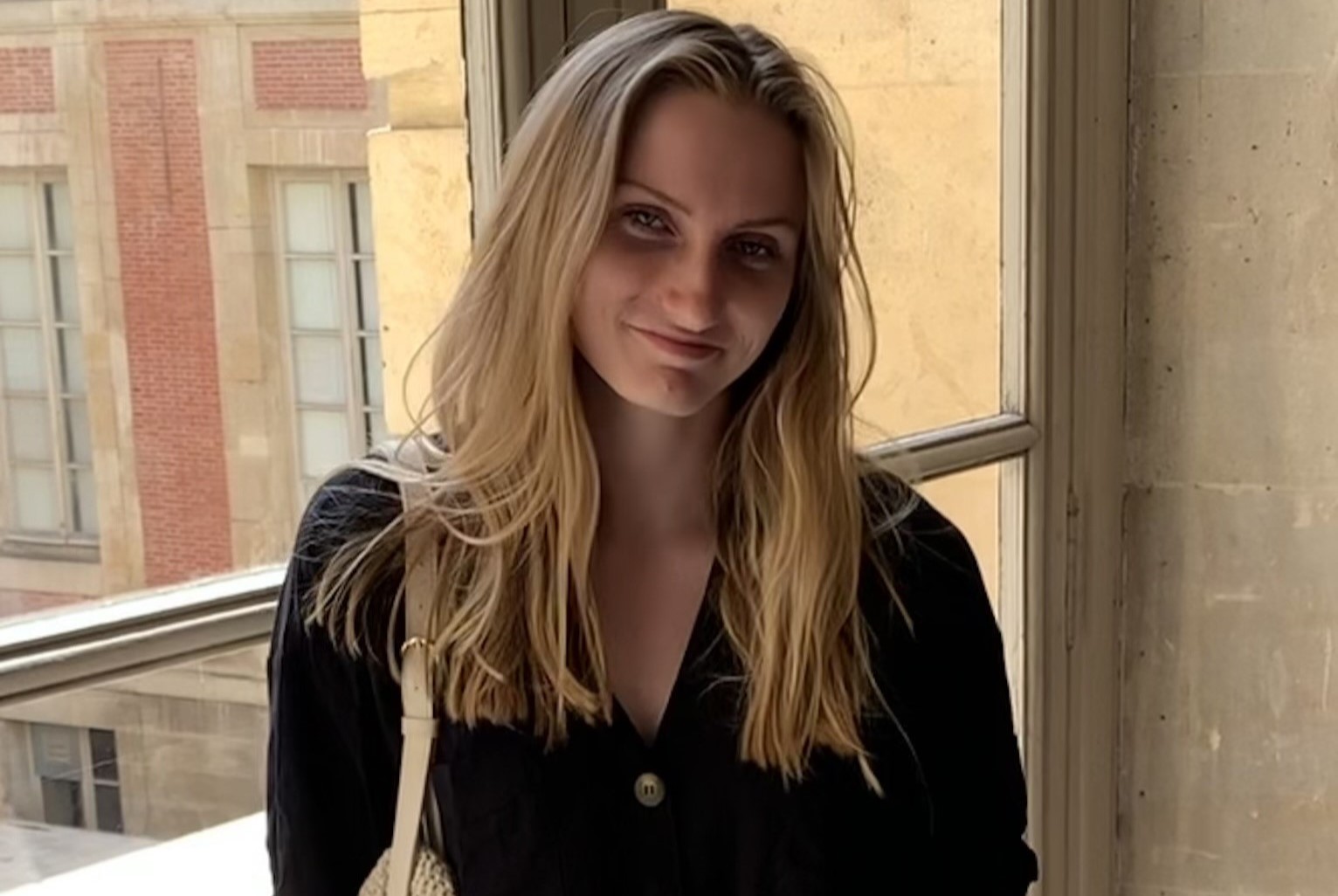 Photo of Jenna Wainwright in front of a window. She is wearing a black shirt with buttons and jeans, with a white bag around her right shoulder.