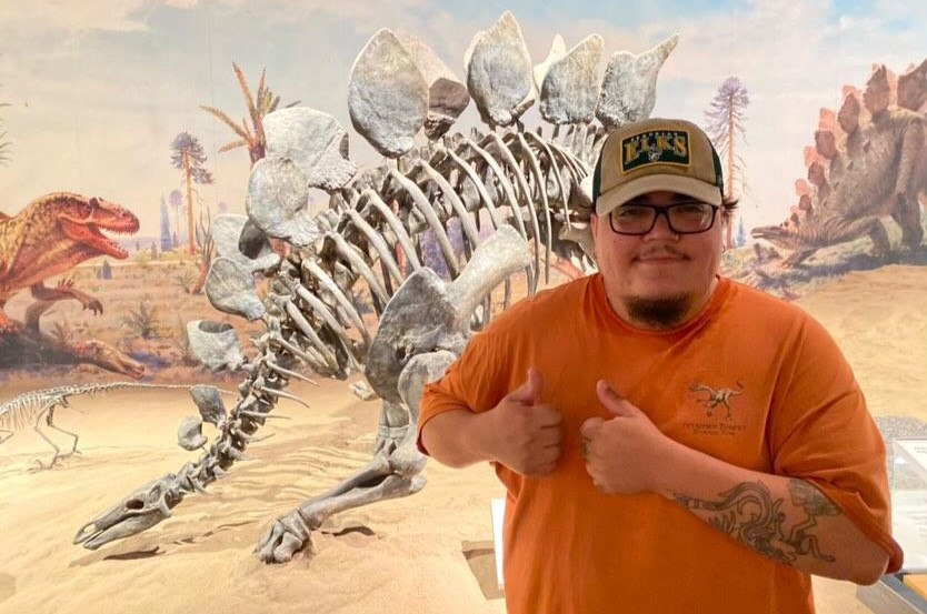 Jeremy Rascon wearing an orange t-shirt and beige hat in front of a stegosaurus skeleton. He is wearing black framed glasses and giving a double thumbs up. 