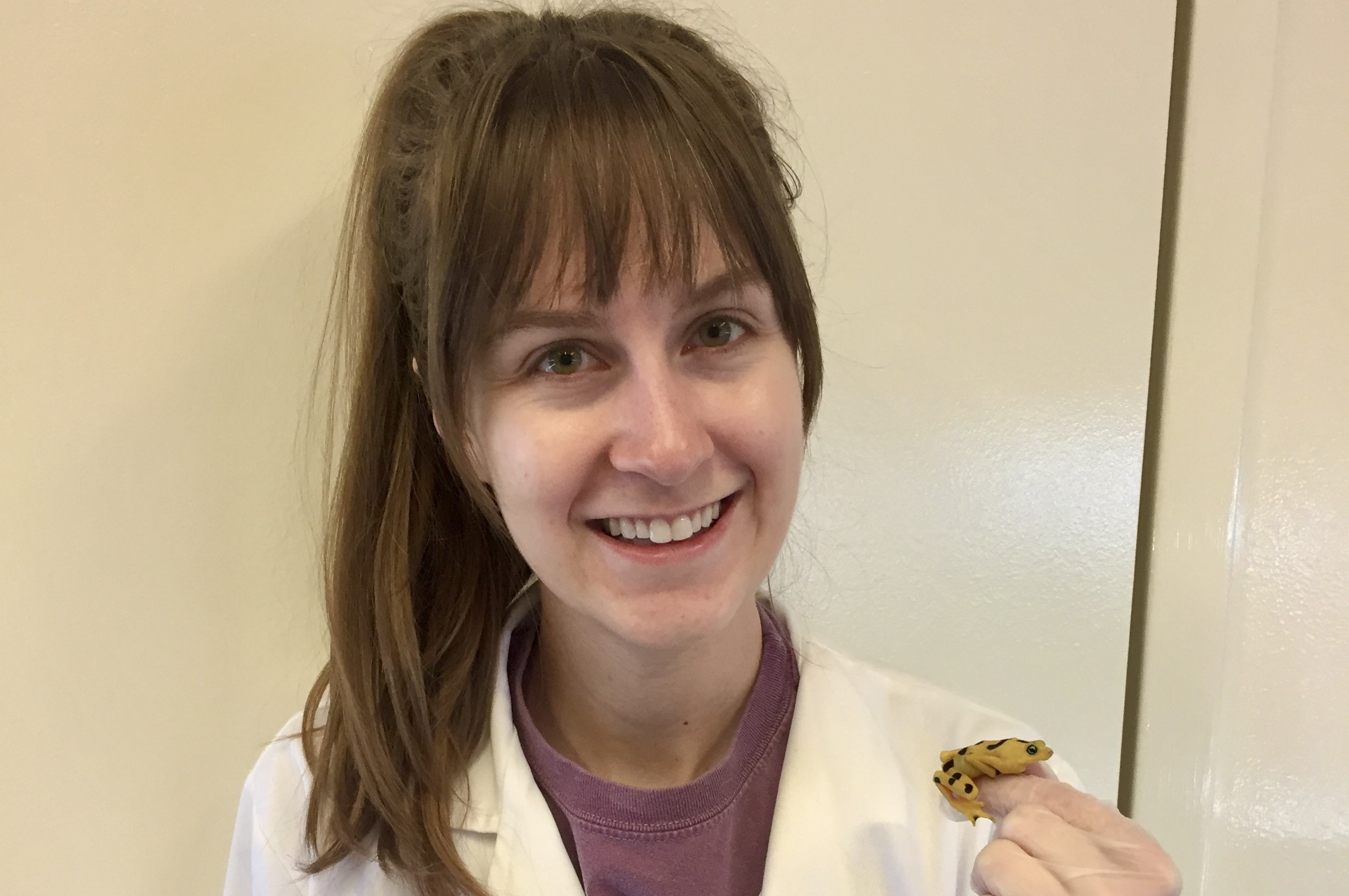 Jordan Gass smiling in a white lab coat, holding a yellow frog with black spots in her left hand.