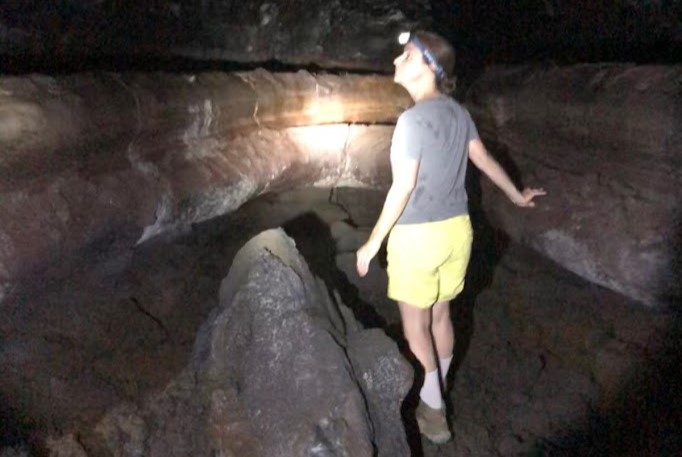 Dr. Kathryn Hanley walking away from camera in a lava tube. She is wearing a headlamp that is illuminating her surroundings, has her right arm out-streched, and is looking up and to her left.
