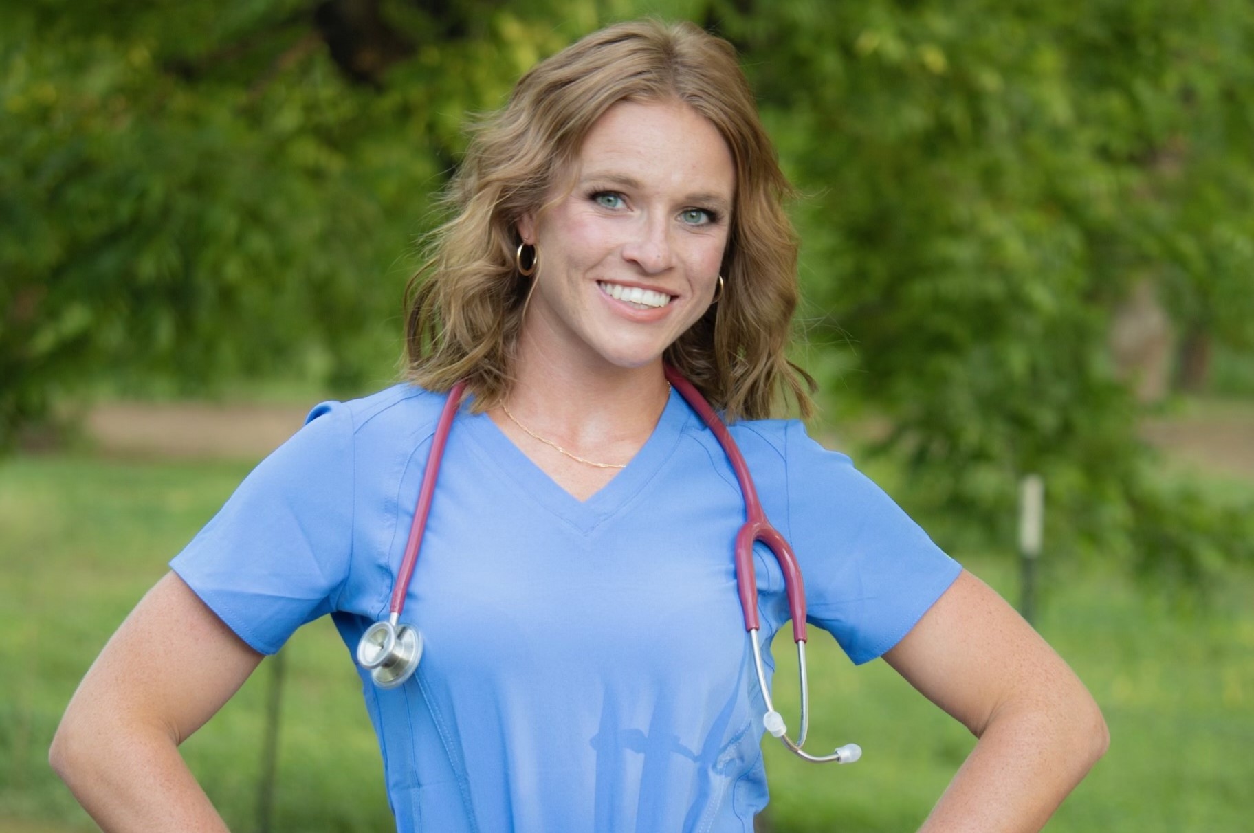 Madelin Whelpley wearing bright blue scrubs and smiling with her hands on her hips. A stethoscope hangs around her neck as she stand in front of some green trees.