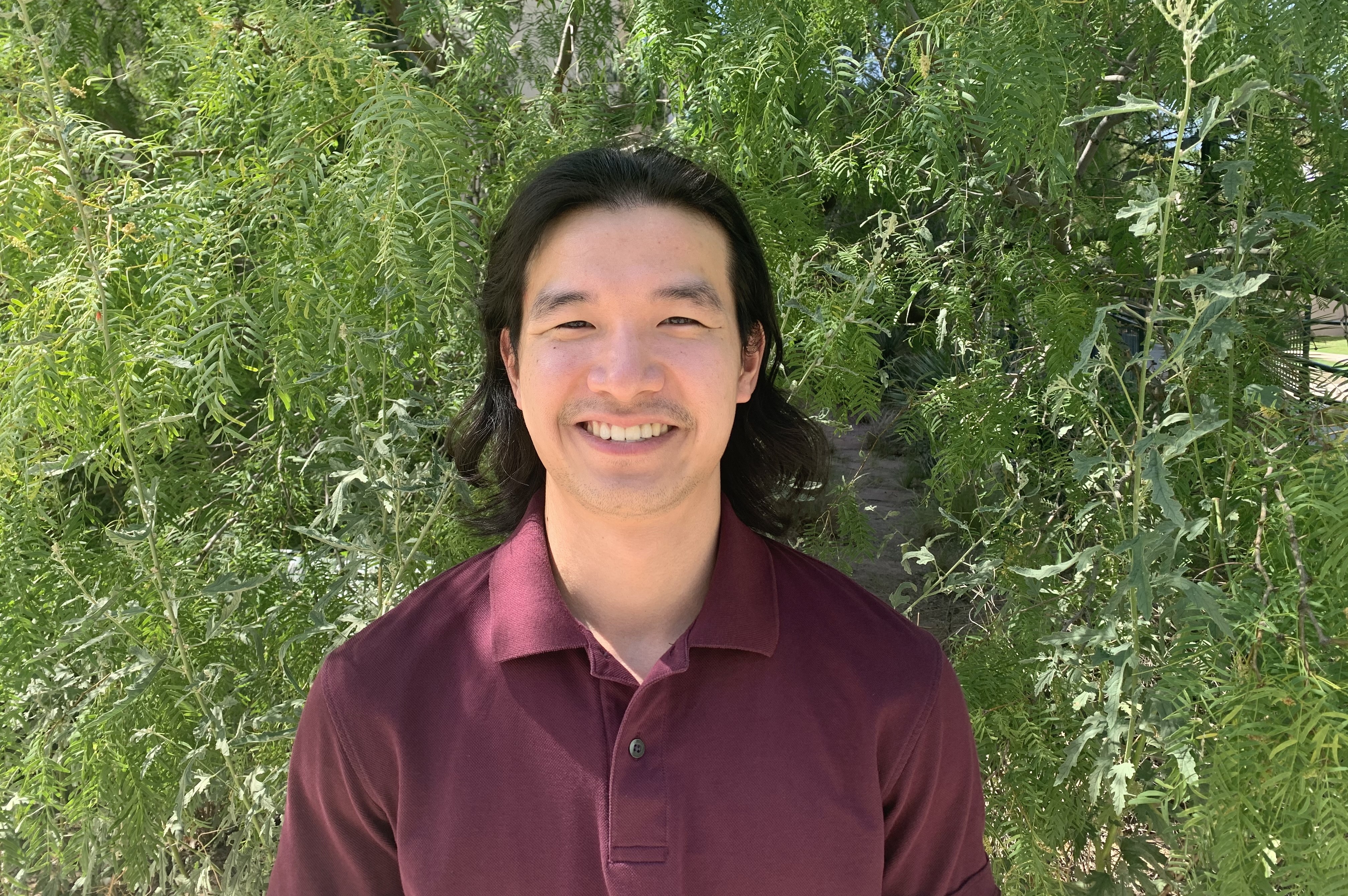 Lawrence Zhou wearing a crimson shirt, smiling in front of green leaves.