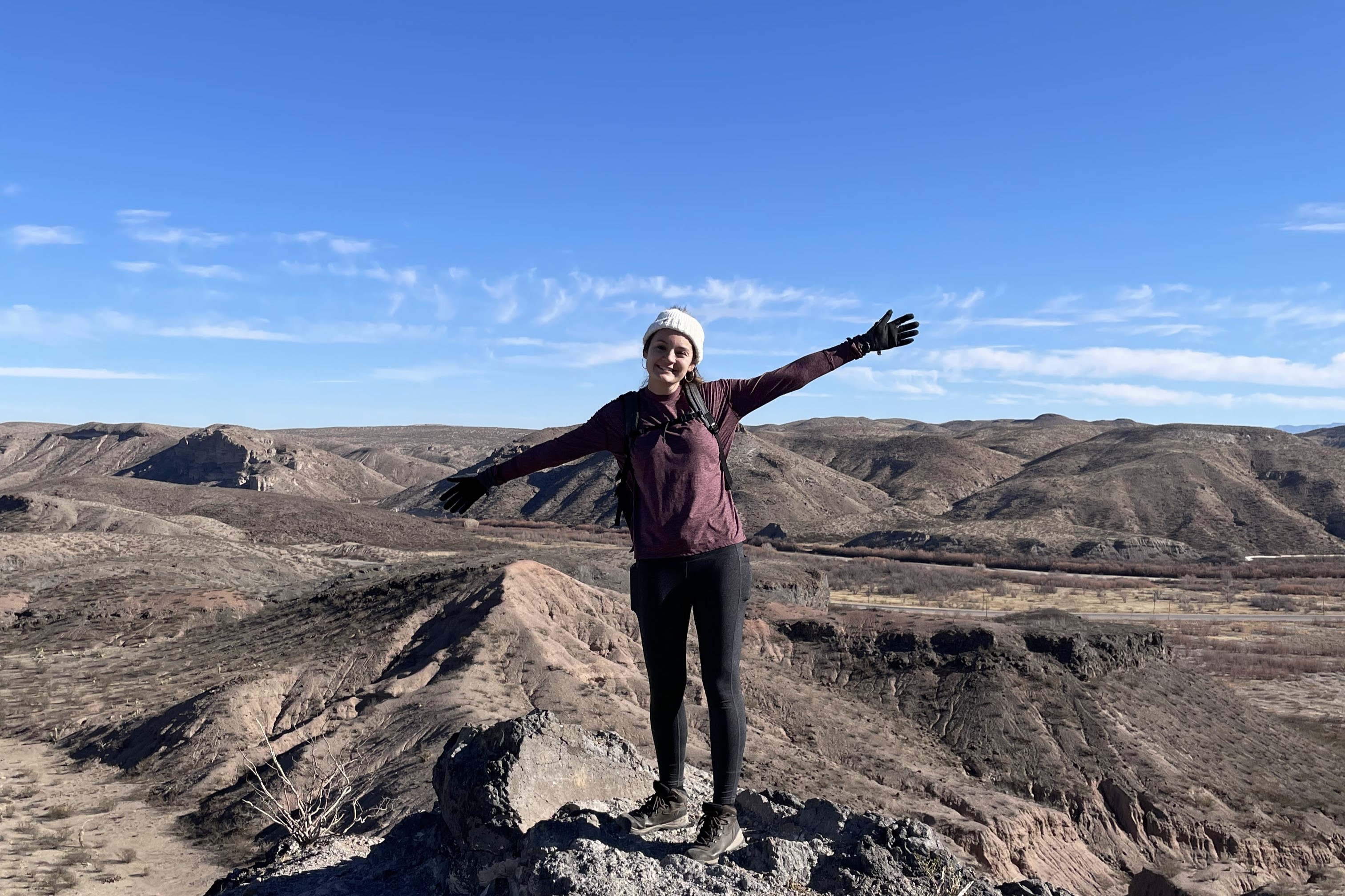 Photo of Marissa Padilla atop some rocks with both arms out-stretched. A clear blue sky fills the top half of the background, while desert hills fill the bottom half of the background.